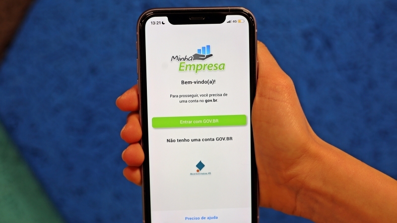 M&atilde;o segura um celular, com um fundo azul. Na tela do celular, est&aacute; o aplicativo Minha Empresa.