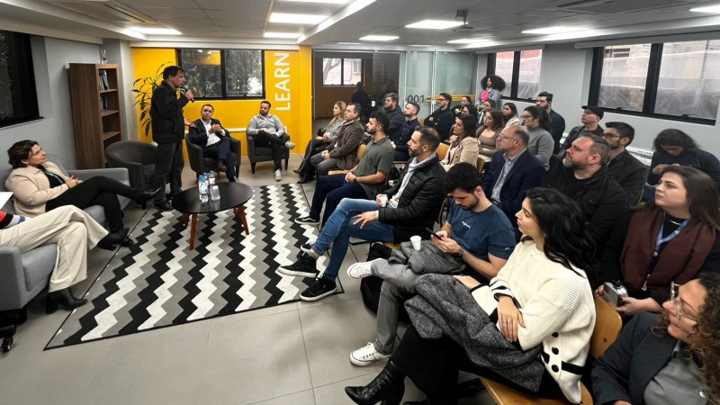 &Agrave; esquerda, um homem fala no microfone, ao lado de dois homens e uma mulher sentados. &Agrave; direita, grupo de 25 pessoas acompanha a fala.