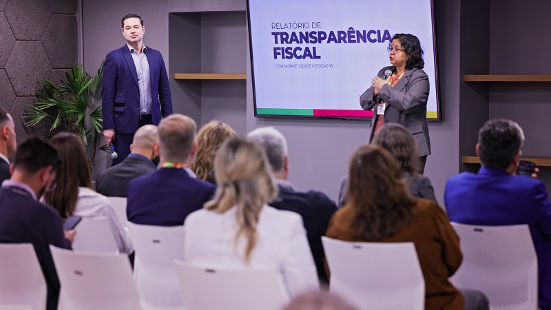 Pricilla Santana e Felipe Bittencourt aparecem em um palco &agrave; frente de um p&uacute;blico. Em um tel&atilde;o, est&aacute; escrito Relat&oacute;rio de Transpar&ecirc;ncia Fiscal. 