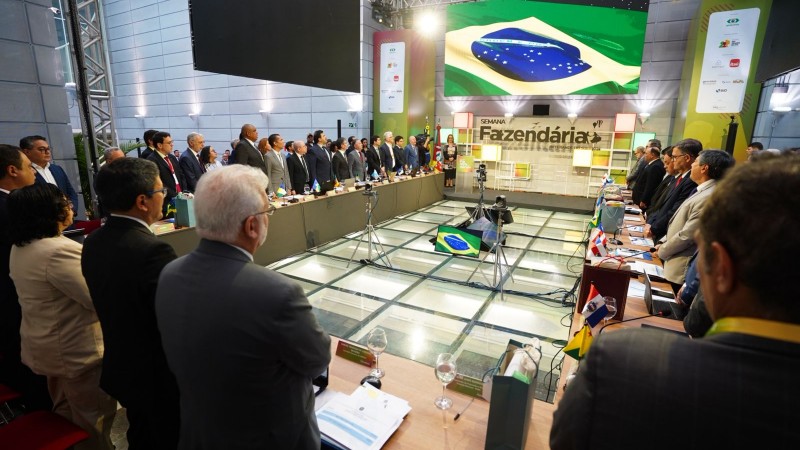 Imagem mostra dezenas de pessoas reunidas em uma mesa em formato de U. Ao fundo, est&aacute; a bandeira do Brasil projetada em um tel&atilde;o e a inscri&ccedil;&atilde;o Semana Fazend&aacute;ria.