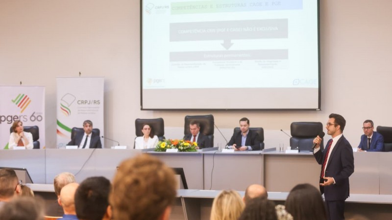 Felipe Reis fala ao microfone. Ao fundo, os componentes de uma mesa de abertura de evento. De costas, &eacute; poss&iacute;vel ver pessoas acompanhando.