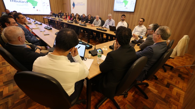 A imagem mostra uma reuni&atilde;o em uma sala de confer&ecirc;ncias ampla, com uma mesa grande em formato retangular ocupando o centro do espa&ccedil;o. Diversas pessoas, homens e mulheres, est&atilde;o sentadas ao redor da mesa, todas voltadas para o centro da discuss&atilde;o. A maioria utiliza notebooks, tablets ou blocos de anota&ccedil;&atilde;o. Ao fundo, h&aacute; duas telas grandes fixadas na parede de madeira clara, exibindo gr&aacute;ficos e mapas relacionados ao tema tribut&aacute;rio. As cadeiras s&atilde;o pretas e girat&oacute;rias, e o piso &eacute; de madeira em padr&atilde;o geom&eacute;trico. O clima transmite um encontro formal, provavelmente t&eacute;cnico ou institucional.