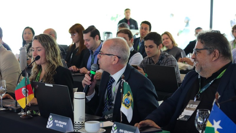 A imagem mostra uma reuni&atilde;o formal com v&aacute;rias pessoas sentadas ao redor de uma grande mesa, todas voltadas para a mesma dire&ccedil;&atilde;o. Em primeiro plano, h&aacute; tr&ecirc;s participantes na mesa principal. Ao centro, um homem de terno segura um microfone enquanto fala; &agrave; sua frente h&aacute; uma pequena bandeira estadual e um notebook aberto. &Agrave; esquerda dele, uma mulher tamb&eacute;m est&aacute; com um microfone pr&oacute;ximo &agrave; boca e utiliza um laptop. &Agrave; direita, outro homem observa a fala com aten&ccedil;&atilde;o. Todos usam crach&aacute;s e est&atilde;o em um ambiente iluminado, com outras pessoas ao fundo acompanhando a reuni&atilde;o, algumas anotando e outras usando computadores. Bandeiras estaduais est&atilde;o posicionadas sobre a mesa, indicando a representa&ccedil;&atilde;o de diferentes regi&otilde;es.