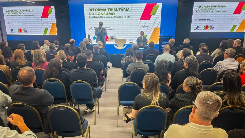 No fundo da foto, em um palco, Ricardo Neves fala ao microfone. Atr&aacute;s dele, &eacute; poss&iacute;vel ver uma apresenta&ccedil;&atilde;o com o t&iacute;tulo Reforma Tribut&aacute;ria do Consumo. &Agrave; frente, dezenas de pessoas acompanham. 