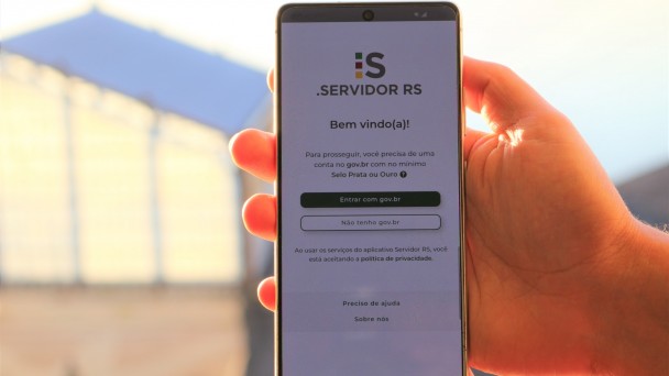 Uma m&atilde;o segura um smartphone na posi&ccedil;&atilde;o vertical, ao ar livre, com a luz do sol iluminando o fundo. Na tela do celular aparece a p&aacute;gina inicial do aplicativo Servidor RS. No topo est&aacute; o logotipo &ldquo;i:s Servidor RS&rdquo; e a mensagem &ldquo;Bem-vindo(a)!&rdquo;. Abaixo, l&ecirc;-se que, para prosseguir, &eacute; necess&aacute;rio ter uma conta no gov.br com n&iacute;vel Selo Prata ou Ouro. H&aacute; dois bot&otilde;es: &ldquo;Entrar com gov.br&rdquo; (em destaque) e &ldquo;N&atilde;o tenho gov.br&rdquo;. Na parte inferior, h&aacute; um aviso sobre pol&iacute;tica de privacidade e um link &ldquo;Preciso de ajuda&rdquo;. O fundo da imagem &eacute; desfocado, sugerindo um ambiente externo com uma estrutura clara ao longe.