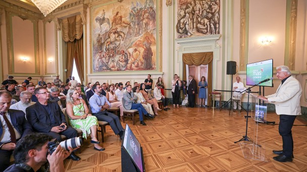 Na imagem, v&ecirc;-se um grande sal&atilde;o de estilo cl&aacute;ssico, com paredes altas em tom claro, detalhes dourados, lustres e grandes pain&eacute;is art&iacute;sticos pintados nas paredes. O piso &eacute; de madeira, com desenho geom&eacute;trico.

&Agrave; direita, um homem em p&eacute; fala ao p&uacute;blico atr&aacute;s de um p&uacute;lpito transparente, usando microfone. Ele veste roupa clara e est&aacute; voltado para a plateia. Ao lado dele h&aacute; uma tela de apresenta&ccedil;&atilde;o apoiada em um suporte, exibindo um slide com fundo colorido e texto.

&Agrave; esquerda e ao centro, h&aacute; v&aacute;rias pessoas sentadas em cadeiras organizadas em fileiras, todas voltadas para o palestrante. O p&uacute;blico &eacute; composto por homens e mulheres de diferentes idades, muitos usando roupas sociais ou casuais. Algumas pessoas observam atentamente, enquanto outras seguram celulares ou c&acirc;meras.

Na parte frontal esquerda, um fot&oacute;grafo est&aacute; agachado, apontando uma c&acirc;mera profissional em dire&ccedil;&atilde;o ao palestrante. Ao fundo, pr&oacute;ximo &agrave;s paredes, h&aacute; mais pessoas em p&eacute;, acompanhando o evento. O ambiente transmite a ideia de uma apresenta&ccedil;&atilde;o formal, como uma coletiva de imprensa, palestra ou an&uacute;ncio oficial em um pr&eacute;dio hist&oacute;rico.