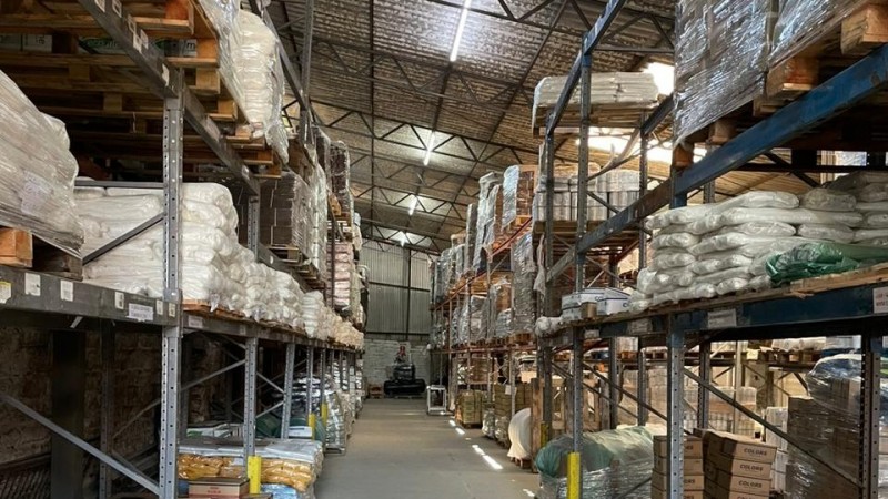 Interior de um grande armaz&eacute;m log&iacute;stico com p&eacute;-direito alto e estrutura met&aacute;lica no teto. H&aacute; corredores largos entre estantes industriais altas, feitas de metal, que v&atilde;o do ch&atilde;o at&eacute; pr&oacute;ximo do teto. As prateleiras est&atilde;o carregadas com diversos pallets e grandes sacos brancos empilhados e embalados em pl&aacute;stico. Tamb&eacute;m h&aacute; caixas e fardos organizados nas estruturas. No centro do corredor, ao fundo, aparece uma empilhadeira utilizada para movimenta&ccedil;&atilde;o de cargas. A ilumina&ccedil;&atilde;o vem de lumin&aacute;rias no teto e de algumas aberturas laterais, criando um ambiente claro e organizado, t&iacute;pico de um dep&oacute;sito de armazenamento de mercadorias.