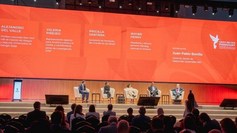 Fotografia de um painel de debate realizado em um grande audit&oacute;rio. No palco, cinco pessoas est&atilde;o sentadas em poltronas claras dispostas em semic&iacute;rculo, participando de uma conversa mediada. &Agrave; frente delas h&aacute; pequenas mesas de apoio e monitores no ch&atilde;o. Ao fundo, um enorme tel&atilde;o laranja ocupa toda a parede do palco, exibindo os nomes dos participantes e da pessoa respons&aacute;vel pela media&ccedil;&atilde;o do debate. &Agrave; direita do tel&atilde;o aparece o logotipo do evento &ldquo;Paraguay&rdquo;.