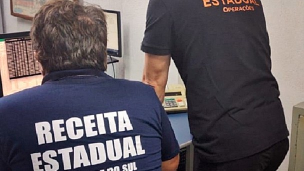 A imagem mostra dois homens em um ambiente de escrit&oacute;rio, vistos de costas, trabalhando diante de um computador. Um deles est&aacute; sentado em uma cadeira e usa uma camiseta azul-marinho com a inscri&ccedil;&atilde;o &ldquo;Receita Estadual &ndash; Rio Grande do Sul&rdquo; nas costas. Ao lado, outro homem est&aacute; em p&eacute;, levemente inclinado em dire&ccedil;&atilde;o &agrave; tela, vestindo uma camiseta preta com a inscri&ccedil;&atilde;o &ldquo;Receita Estadual &ndash; Opera&ccedil;&otilde;es&rdquo; em letras laranja. Ambos parecem concentrados no monitor &agrave; frente. Na parede, ao lado do computador, h&aacute; um mapa e alguns materiais fixados, sugerindo um ambiente de trabalho administrativo ligado &agrave; &aacute;rea fiscal.