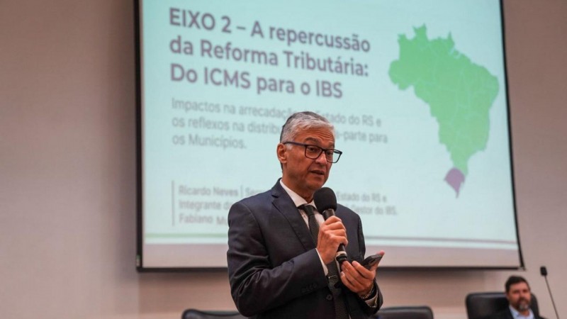 Ricardo Neves Pereira fala ao microfone. Ao fundo, um tel&atilde;o exibe a mensagem "A repercuss&atilde;o da Reforma Tribut&aacute;ria: do ICMS para o IBS". 