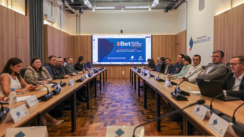 Sala de reuni&otilde;es ampla, com paredes revestidas em madeira clara e ilumina&ccedil;&atilde;o embutida no teto. No centro, duas fileiras de mesas compridas formam um corredor, com cerca de vinte pessoas sentadas, distribu&iacute;das dos dois lados, todas voltadas para o fundo da sala.

Os participantes s&atilde;o homens e mulheres adultos, vestidos de forma formal ou semi-formal, muitos com ternos, blazers e camisas sociais. &Agrave; frente de cada pessoa h&aacute; um microfone, garrafas de &aacute;gua e identificadores de mesa, indicando um encontro institucional ou t&eacute;cnico.

Ao fundo, h&aacute; uma grande tela de proje&ccedil;&atilde;o exibindo uma apresenta&ccedil;&atilde;o com o t&iacute;tulo &ldquo;IBET&rdquo; e gr&aacute;ficos em tons de azul, sugerindo um tema relacionado a economia, tecnologia ou dados. No piso, o ch&atilde;o &eacute; de madeira em padr&atilde;o geom&eacute;trico.

O ambiente transmite um clima de reuni&atilde;o oficial, possivelmente um semin&aacute;rio, workshop ou encontro de trabalho entre representantes de diferentes institui&ccedil;&otilde;es.