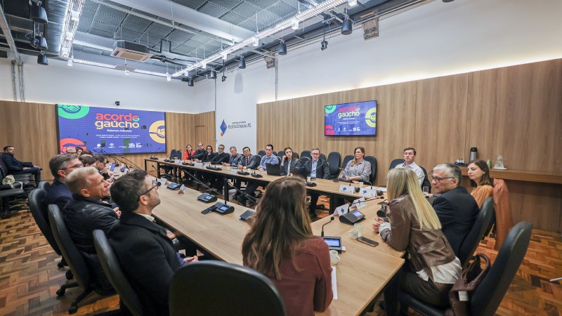 Imagem de uma sala de reuni&otilde;es ampla e bem iluminada, com uma grande mesa retangular de madeira ocupando o centro. Ao redor da mesa, h&aacute; diversas pessoas sentadas, homens e mulheres, vestindo roupas formais e de neg&oacute;cios, participando de um encontro ou apresenta&ccedil;&atilde;o. A maioria est&aacute; voltada para o centro da mesa ou para um dos lados da sala, indicando aten&ccedil;&atilde;o a quem fala.

Sobre a mesa, h&aacute; microfones individuais, notebooks, celulares e alguns pap&eacute;is, sugerindo uma reuni&atilde;o institucional ou coletiva de trabalho. No fundo da sala, duas telas exibem uma apresenta&ccedil;&atilde;o com o t&iacute;tulo &ldquo;Acordo Ga&uacute;cho&rdquo;, acompanhada de elementos gr&aacute;ficos coloridos.

O ambiente possui paredes com revestimento em madeira, ilumina&ccedil;&atilde;o embutida no teto e estrutura aparente, conferindo um aspecto moderno e corporativo. Algumas pessoas parecem estar falando enquanto outras escutam atentamente.