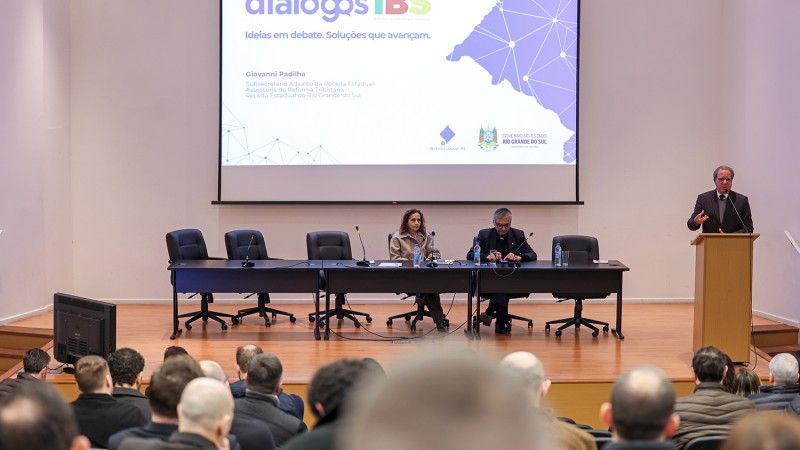 Fotografia de um audit&oacute;rio durante uma palestra ou evento institucional. Em primeiro plano, aparecem as cabe&ccedil;as e os ombros de dezenas de pessoas sentadas na plateia, voltadas para o palco. Ao centro, h&aacute; uma mesa longa de reuni&otilde;es com v&aacute;rias cadeiras. Duas pessoas est&atilde;o sentadas atr&aacute;s da mesa, enquanto um homem fala em p&eacute; em um p&uacute;lpito localizado &agrave; direita do palco. Ao fundo, um grande tel&atilde;o exibe uma apresenta&ccedil;&atilde;o com o t&iacute;tulo &ldquo;Di&aacute;logos IBS&rdquo; e elementos gr&aacute;ficos em tons de azul e roxo. O ambiente &eacute; amplo, com paredes claras, ilumina&ccedil;&atilde;o uniforme e aspecto formal, caracter&iacute;stico de um semin&aacute;rio ou confer&ecirc;ncia.