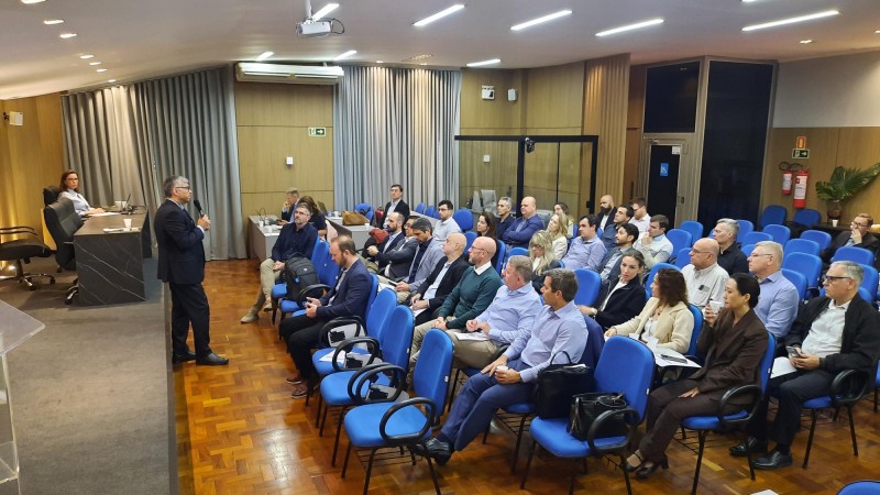 A foto mostra uma pessoa de p&eacute; falando ao microfone para cerca de 30 pessoas sentadas