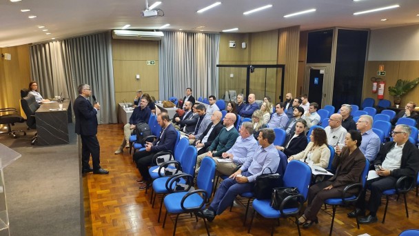 A foto mostra uma pessoa de p&eacute; falando ao microfone para cerca de 30 pessoas sentadas